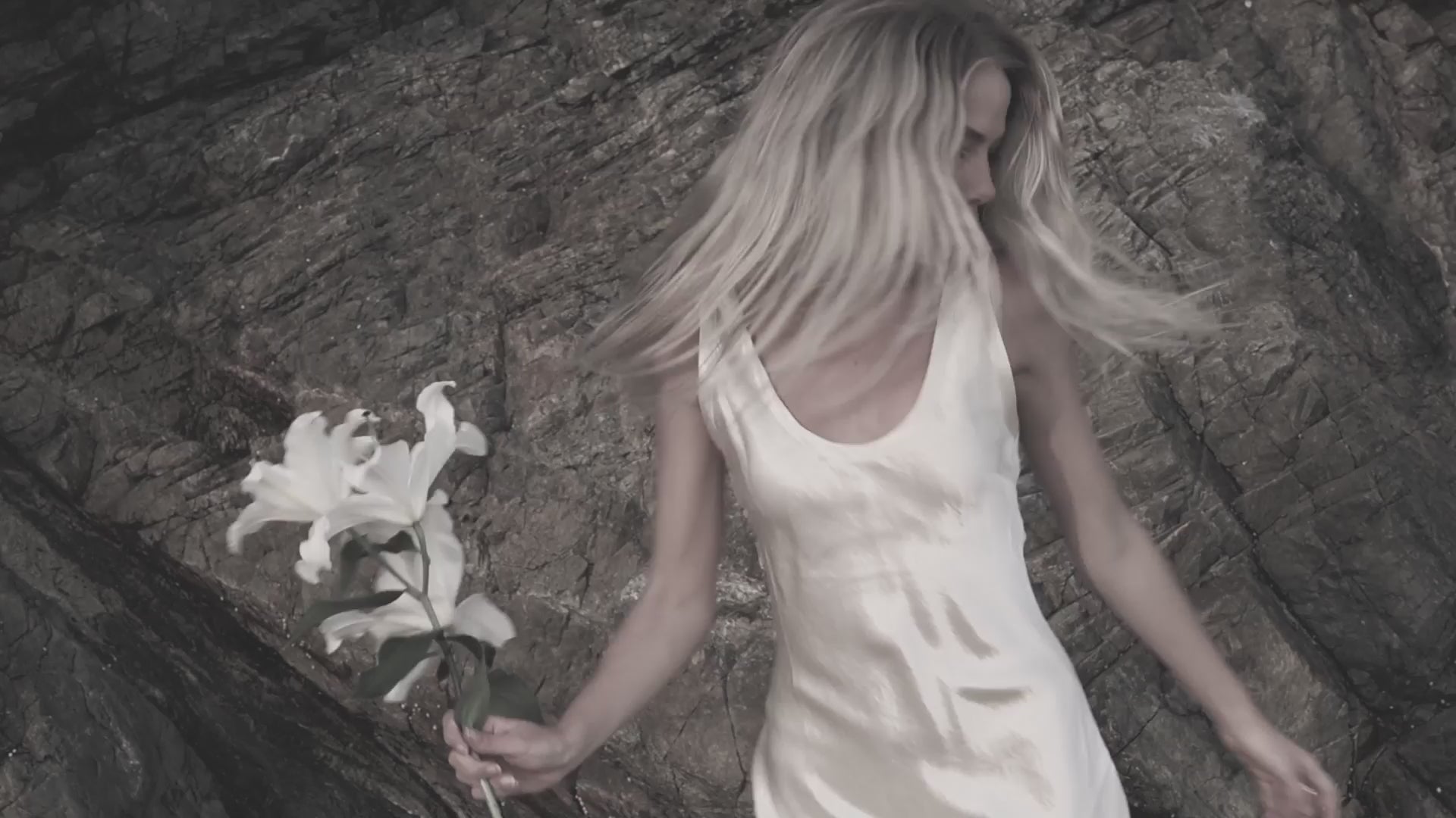 Woman in an elegant white dress holding a white lily against a natural rock background